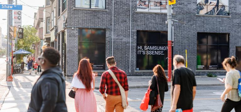 People crossing a street in front of a brick building with posters and advertisements.