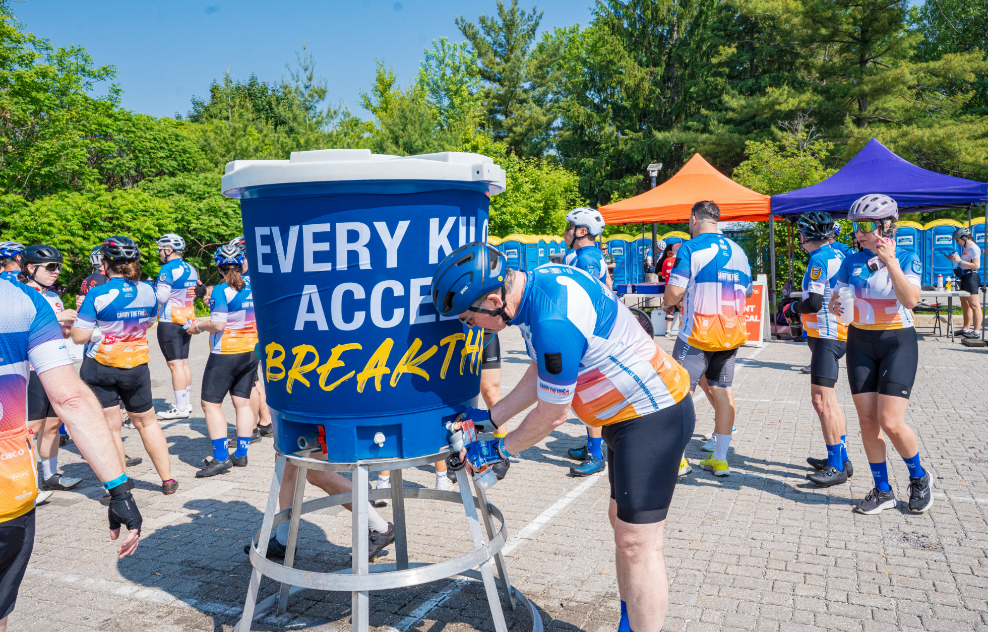 Cyclists wearing matching jerseys gather around a large blue water cooler during a bike event.